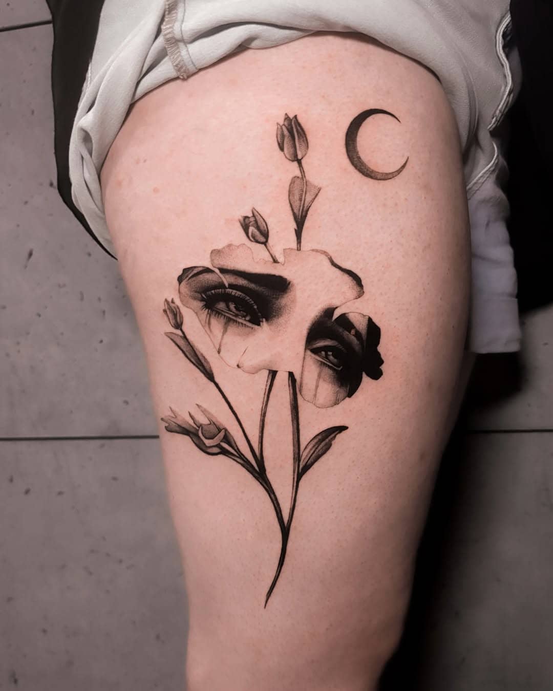 Tatuaż surrealistyczny przedstawiający kobiecą twarz wkomponowaną w kwiaty i księżyc, wykonany na udzie, Nava Sztorm Tattoo Gdańsk.