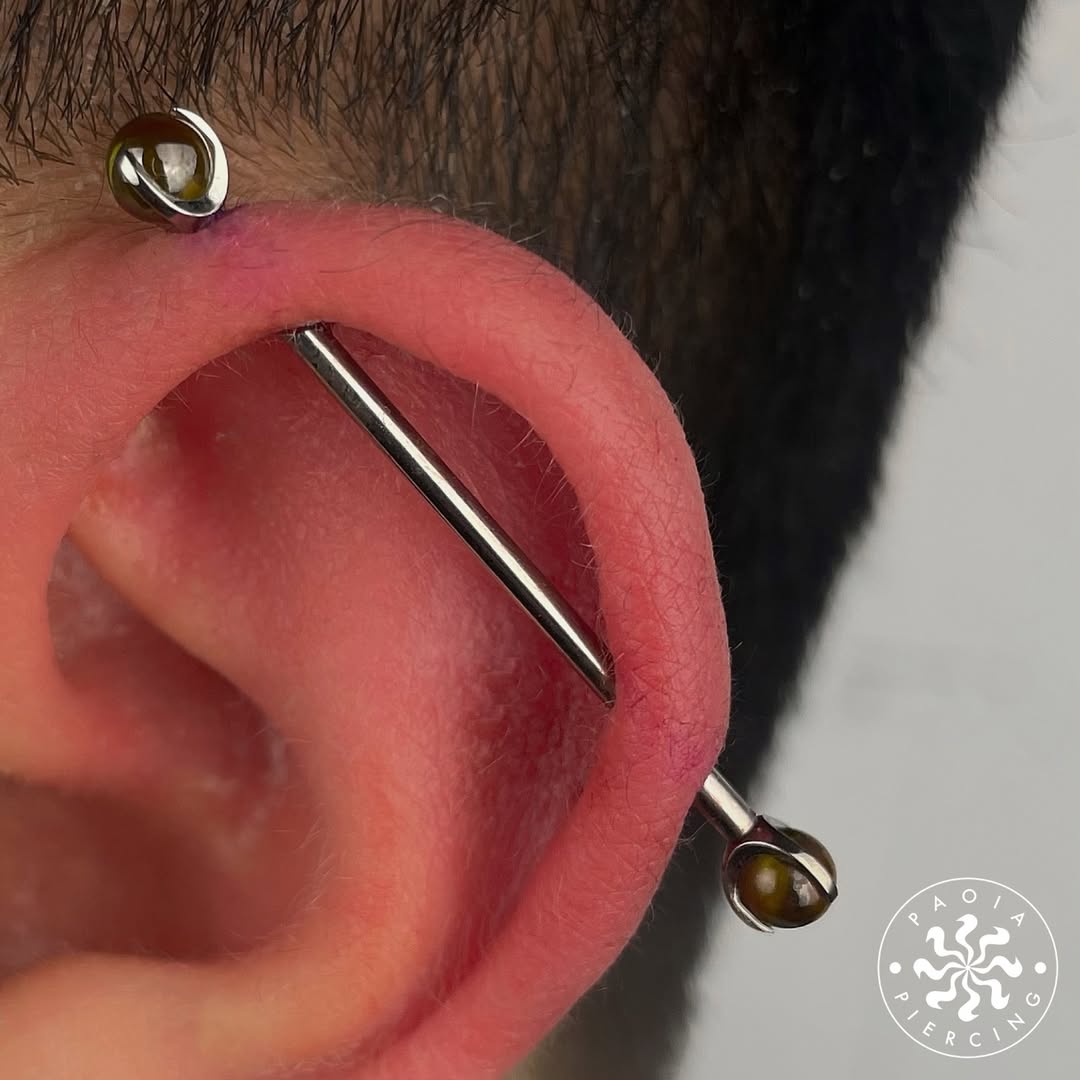 Zbliżenie ucha z industrialnym piercingiem: metalowy pręt przechodzi przez górną część ucha, zakończony dwoma ciemnymi kulkami.