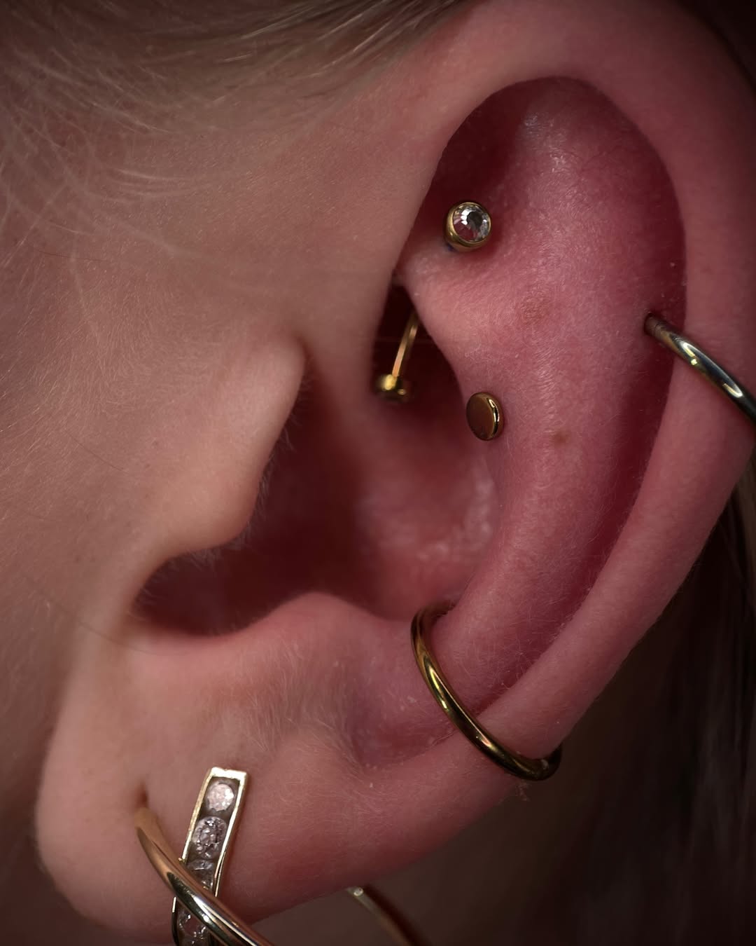 Zbliżenie ucha z ozdobnymi kolczykami w chrząstce i płatku, pokazujące złote elementy i połyskujący kamień; inspiracja do kreatywnego piercingu.