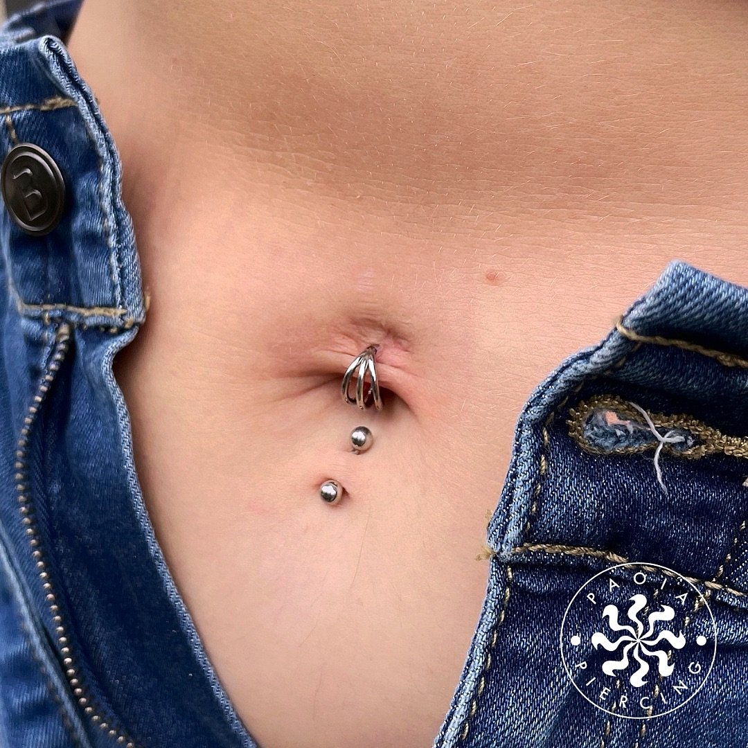 Zbliżenie pępka z piercingiem w kształcie korony oraz dwoma metalowymi kulkami, z fragmentem dżinsów na bokach.