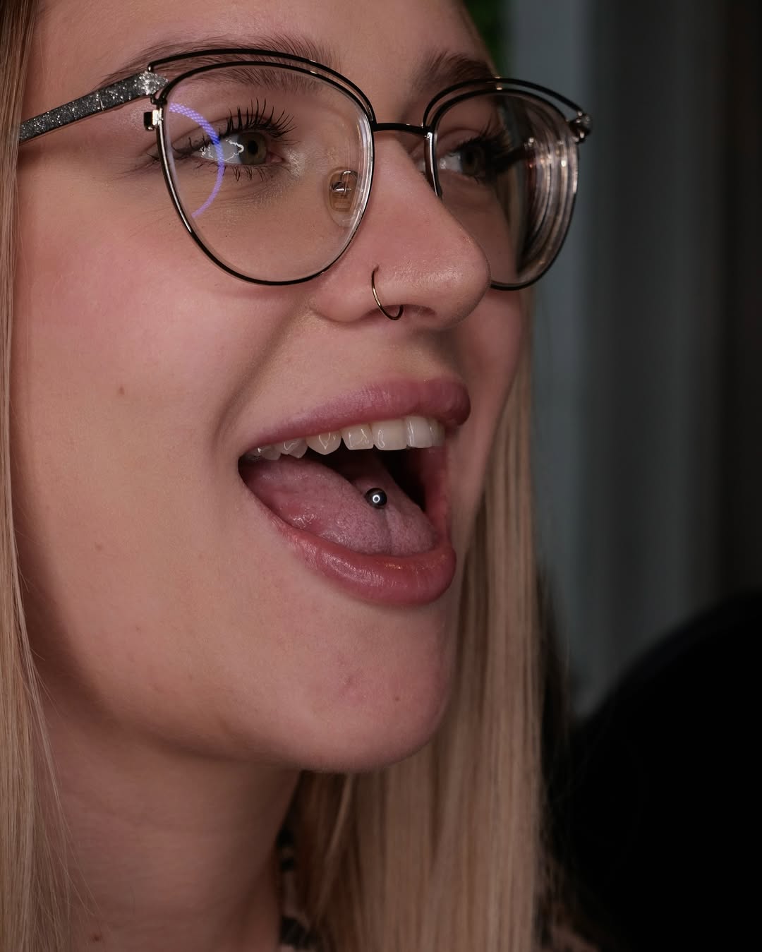 Ujęcie kobiety w okularach z kolczykiem w nosie, która wysuwa język z ciemnym kolczykiem – piercing języka i nosa.